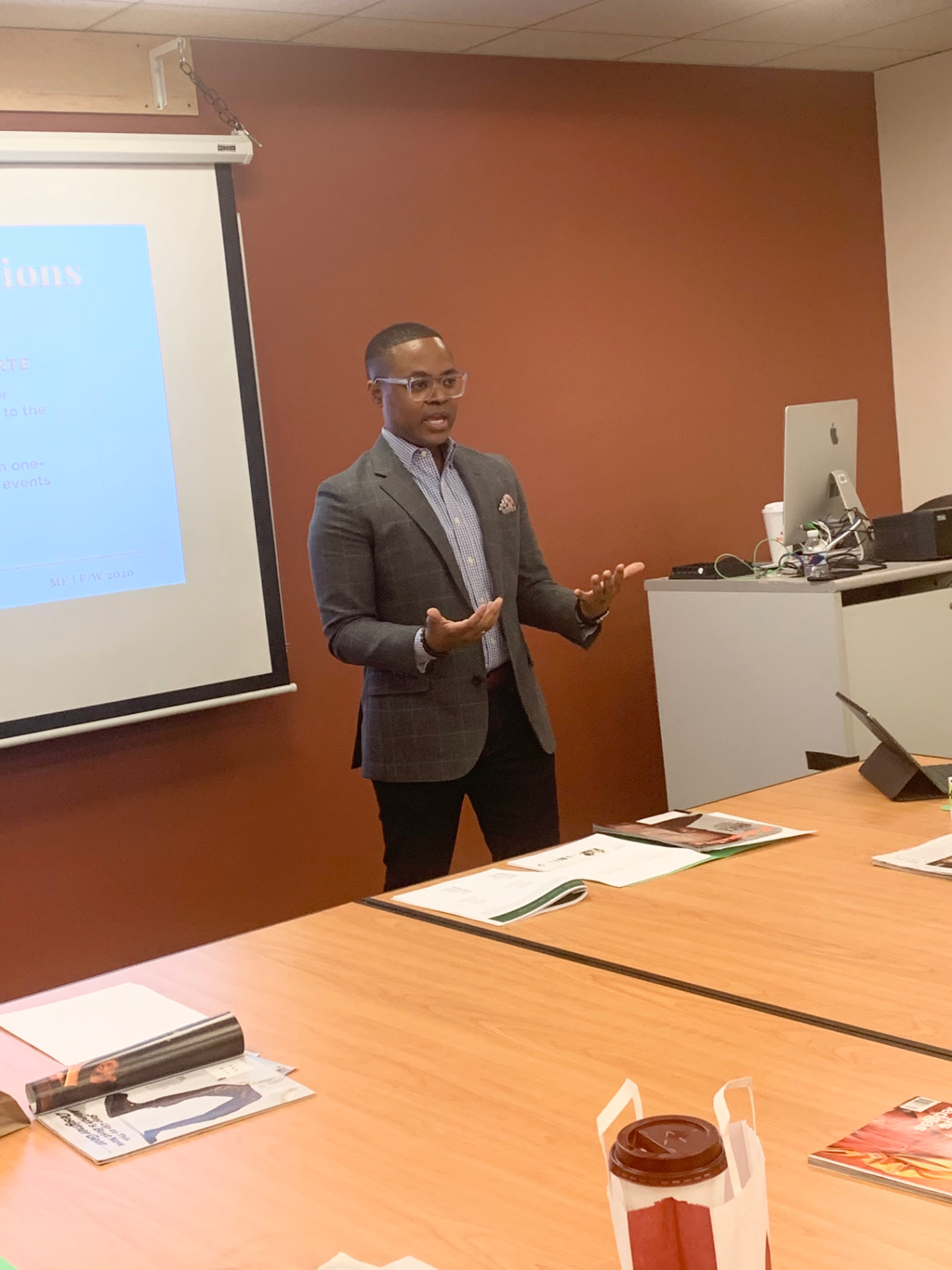 David McKnight presenting in a classroom, gesturing beside a projection screen in a grey checked blazer