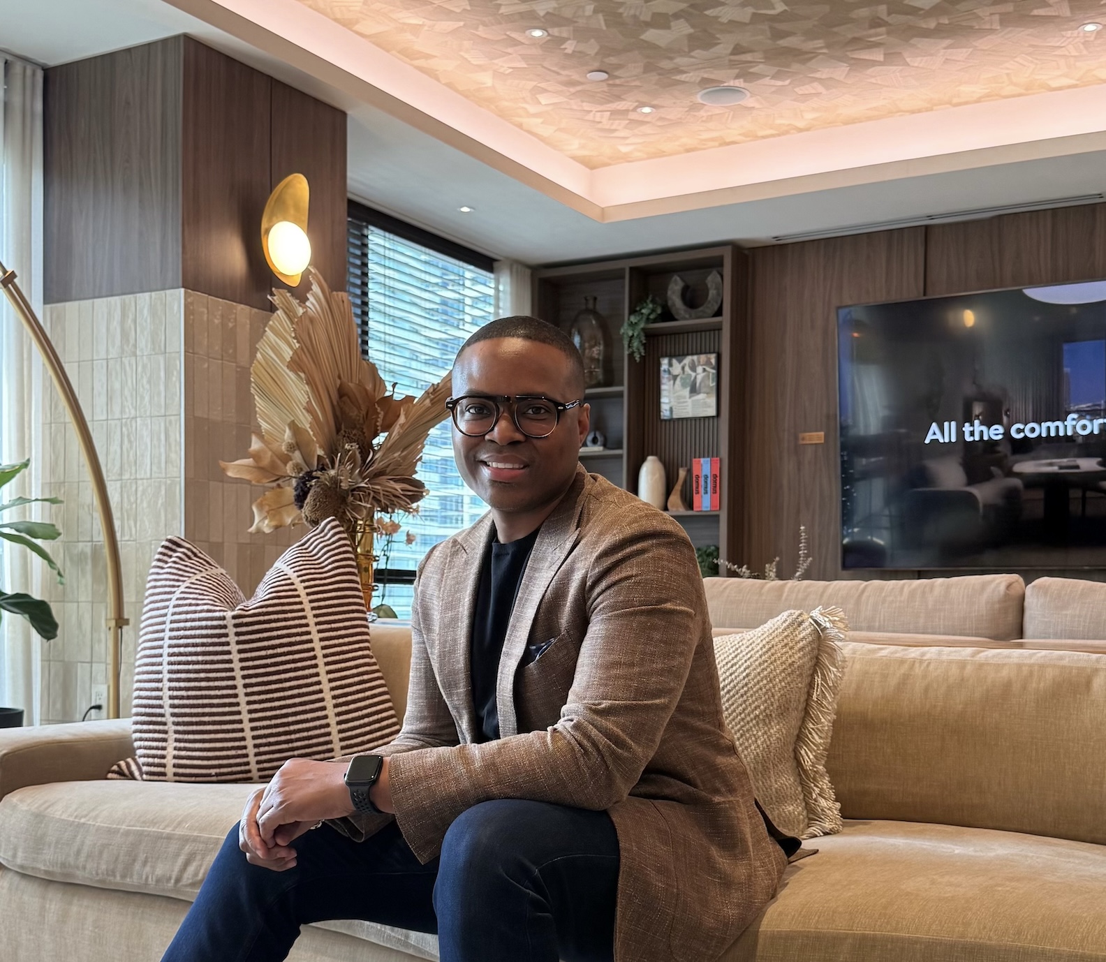 David McKnight seated on a sofa in a tan textured blazer, black crewneck and dark jeans, smiling at the camera